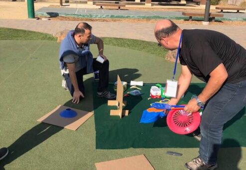 Two participants engaged in a team building 'Build Your Own Mini Golf Course' event, working on constructing a mini golf hole using cardboard, a fan, and other creative materials on a mat laid out on a grassy area outdoors. This activity encourages teamwork and innovation while using everyday objects to create a golf course..