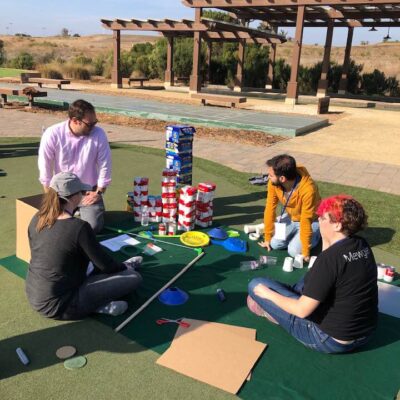 Group of participants sitting on a mat outdoors, collaborating to build a creative mini golf hole using food cans, boxes, and various supplies during a 'Build Your Own Mini Golf Course' team building event. thumbnail