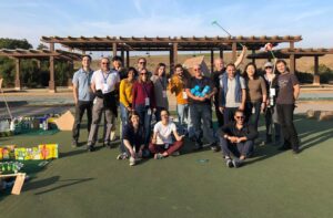 A group of people standing outdoors in a sunny area, posing together after participating in a 'Build Your Own Mini Golf Course' team building event. Some participants hold golf clubs, while others smile for the camera.
