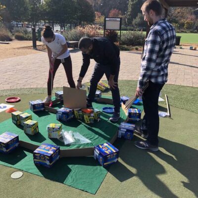 Participants collaborate to build a mini golf hole using boxes of food and other non-perishable items during an outdoor 'Build Your Own Mini Golf Course' team building event. The activity combines creativity, teamwork, and charity, with the food items being donated to a local food pantry afterward. thumbnail