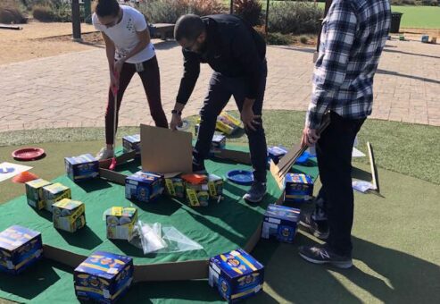 Participants collaborate to build a mini golf hole using boxes of food and other non-perishable items during an outdoor 'Build Your Own Mini Golf Course' team building event. The activity combines creativity, teamwork, and charity, with the food items being donated to a local food pantry afterward.