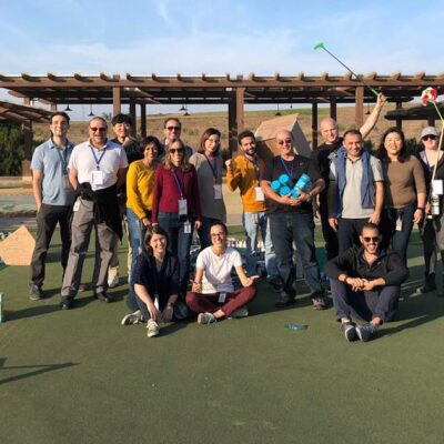 A group of people standing outdoors in a sunny area, posing together after participating in a 'Build Your Own Mini Golf Course' team building event. Some participants hold golf clubs, while others smile for the camera. thumbnail