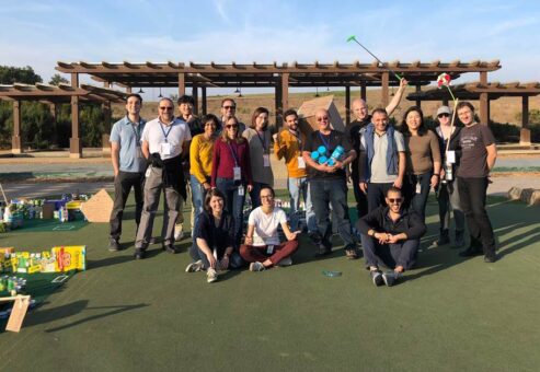 A group of people standing outdoors in a sunny area, posing together after participating in a 'Build Your Own Mini Golf Course' team building event. Some participants hold golf clubs, while others smile for the camera.