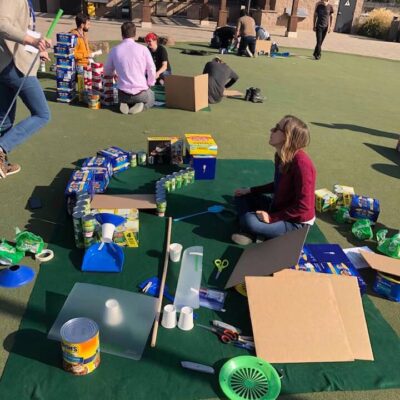 Group of participants sitting on a mat outdoors, collaborating to build a creative mini golf hole using food cans, boxes, and various supplies during a 'Build Your Own Mini Golf Course' team building event. thumbnail