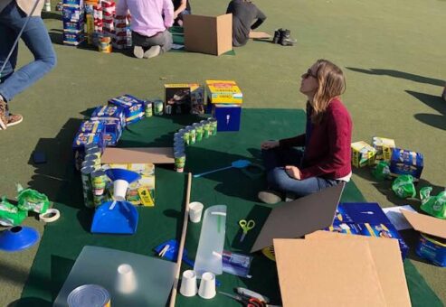 Group of participants sitting on a mat outdoors, collaborating to build a creative mini golf hole using food cans, boxes, and various supplies during a 'Build Your Own Mini Golf Course' team building event.