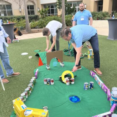 Participants setting up a creative mini golf course using canned goods, cereal boxes, and cardboard during a 'Build Your Own Mini Golf Course' team building event. thumbnail