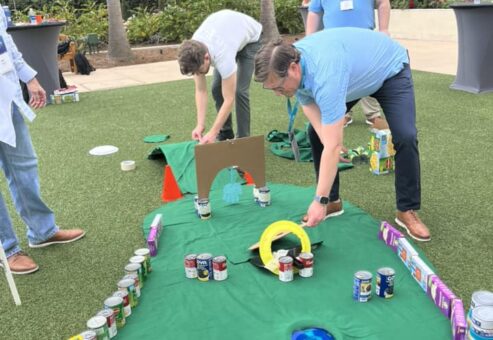 Participants setting up a creative mini golf course using canned goods, cereal boxes, and cardboard during a 'Build Your Own Mini Golf Course' team building event.