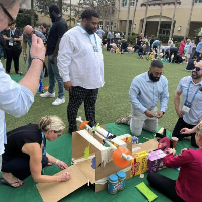 A team of participants collaboratively building a creative mini golf hole using cardboard, canned goods, and food packages during a 'Build Your Own Mini Golf Course' team building event. thumbnail
