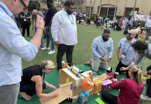 A team of participants collaboratively building a creative mini golf hole using cardboard, canned goods, and food packages during a 'Build Your Own Mini Golf Course' team building event.