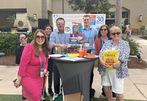 A group of participants posing with canned goods and food packages during a 'Build Your Own Mini Golf Course' team building event. The team stands outdoors, displaying their collection of food items that will be donated to a local charity, combining creativity, teamwork, and a meaningful cause.