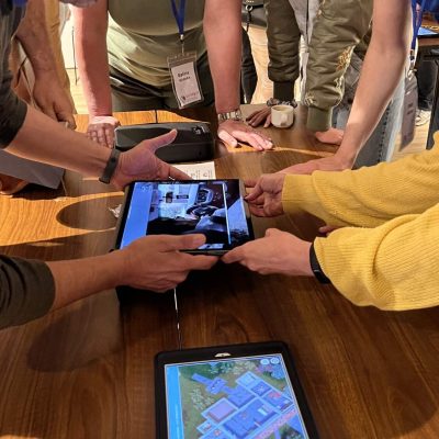 Participants in a 'Corporate CSI' team building event gather around a table, using iPads to examine digital evidence and clues as they work collaboratively to solve the mystery. thumbnail