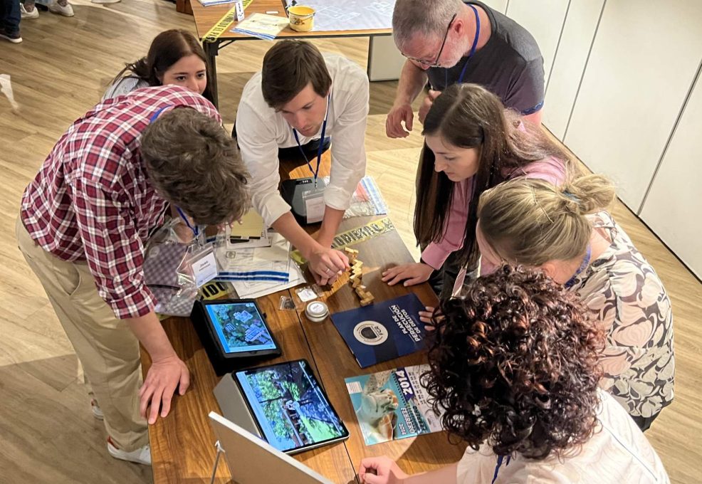 Participants in the 'Corporate CSI' team building challenge gather around a table, focused on solving clues from their detective briefcases and using digital tools to navigate a virtual crime scene.