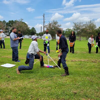 Participants outdoors engaging in a team building exercise with a catapult setup, preparing for a launch while other team members observe and strategize, demonstrating teamwork, collaboration, and competitive spirit in the Catapult to Success activity. thumbnail