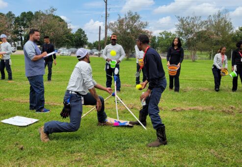 Participants outdoors engaging in a team building exercise with a catapult setup, preparing for a launch while other team members observe and strategize, demonstrating teamwork, collaboration, and competitive spirit in the Catapult to Success activity.