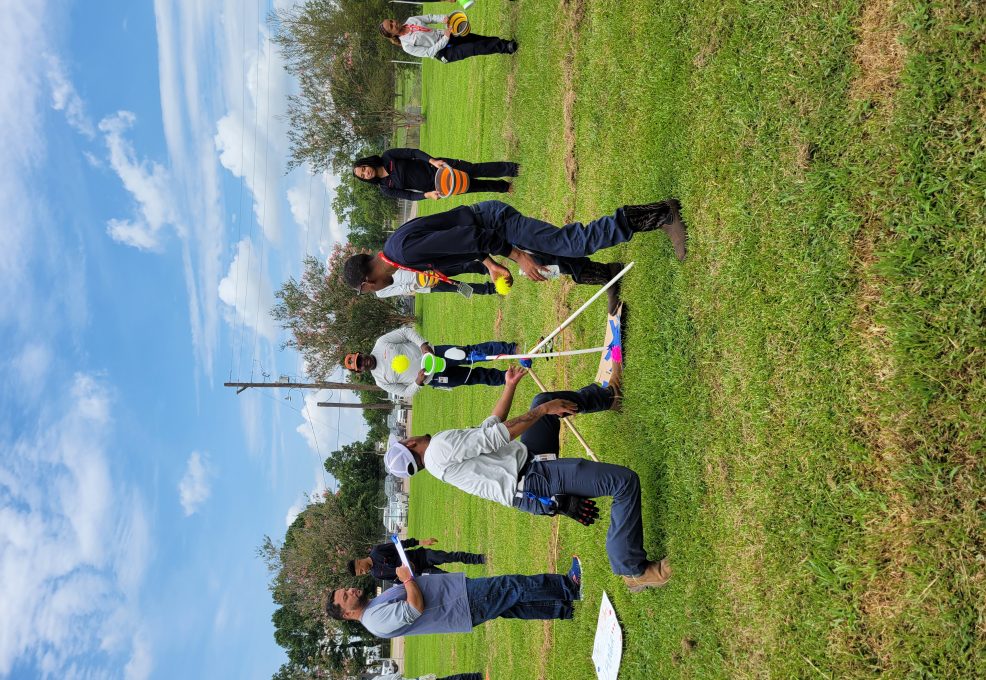 Participants outdoors engaging in a team building exercise with a catapult setup, preparing for a launch while other team members observe and strategize, demonstrating teamwork, collaboration, and competitive spirit in the Catapult to Success activity.