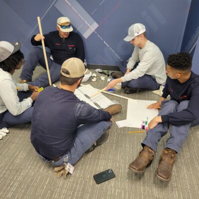 A group of participants sitting on the floor indoors, discussing and planning their catapult design during the Catapult to Success team building activity, emphasizing teamwork, collaboration, and strategic thinking. thumbnail