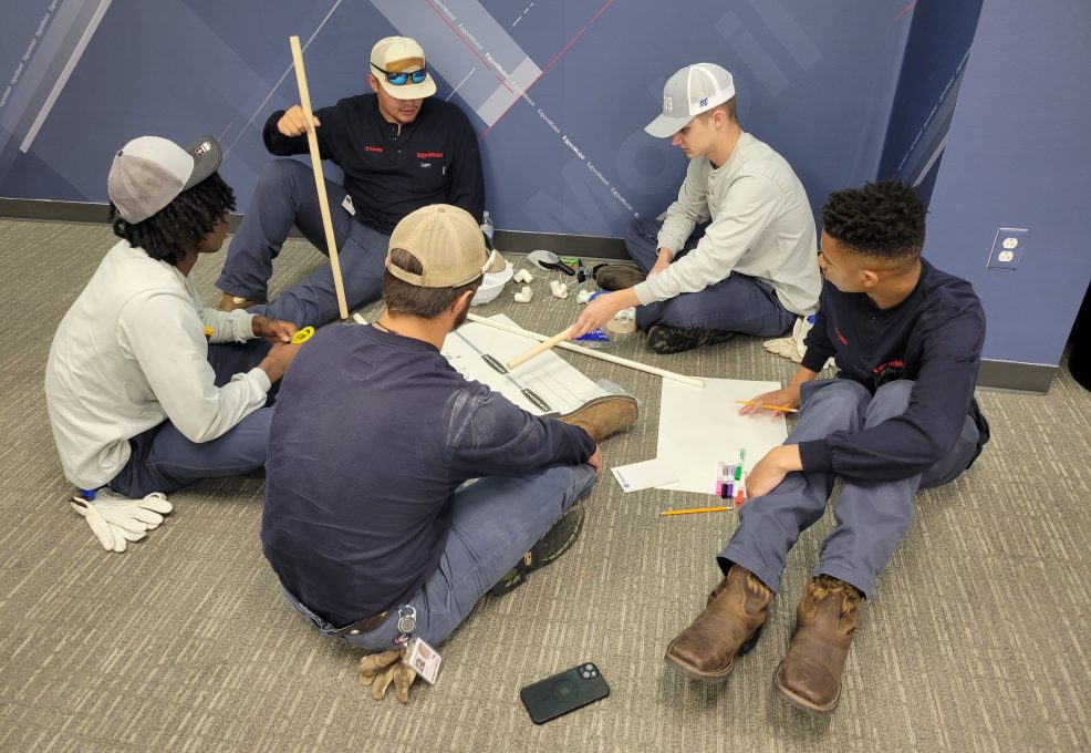 A group of participants sitting on the floor indoors, discussing and planning their catapult design during the Catapult to Success team building activity, emphasizing teamwork, collaboration, and strategic thinking.