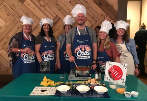 A group of seven people stand behind a table, smiling and wearing chef hats and aprons for a Chili Cook-Off event. They are preparing a chili dish with a pot on a portable stove in front of them. Various ingredients and garnishes are laid out on the table. A sign reads 'We can stop your heart and revive it too!' indicating a playful theme. The group is enjoying a team building culinary event.