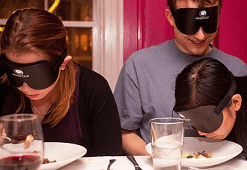 Three people wearing blindfolds while eating at a dining table, participating in a sensory deprivation dining experience to enhance communication and trust.