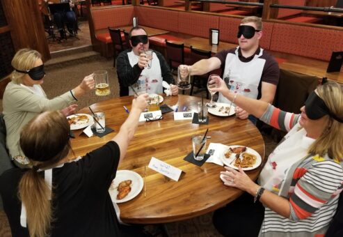 Group of participants blindfolded at a 'Dining In The Dark' team building event, toasting with drinks around a wooden table, enhancing trust and communication skills.