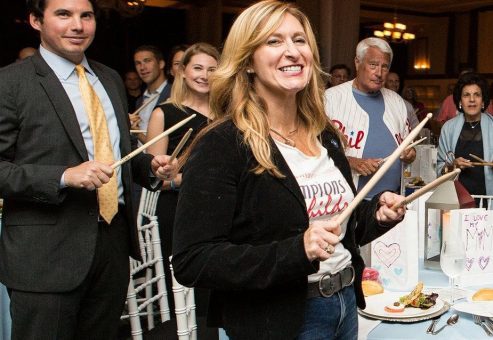 Smiling participants in the Drumming Up A Team event enthusiastically engage in a rhythmic percussion activity, fostering teamwork and energy in a lively group setting.