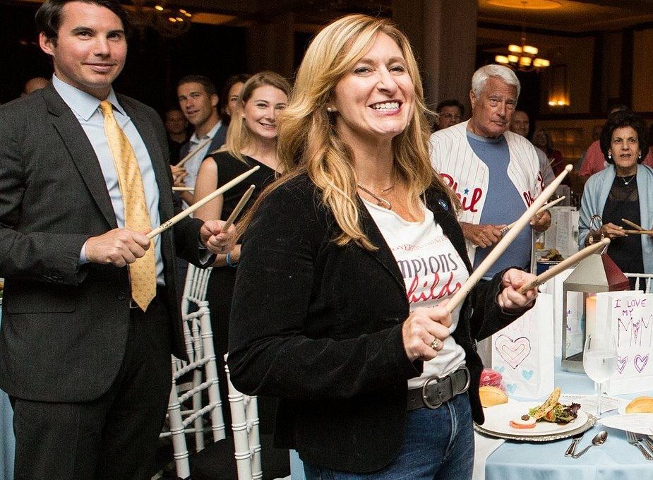 Smiling participants in the Drumming Up A Team event enthusiastically engage in a rhythmic percussion activity, fostering teamwork and energy in a lively group setting.
