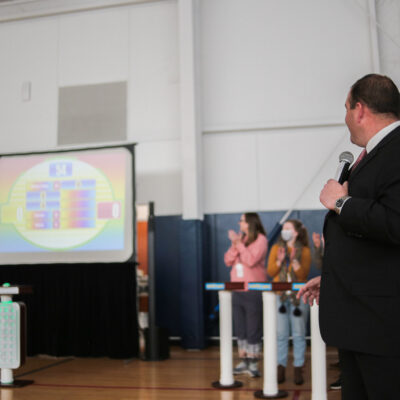 A game show host holding a microphone and looking at a large screen displaying the game board, while participants cheer and clap during the Corporate Feud team building activity in a gymnasium setting.
