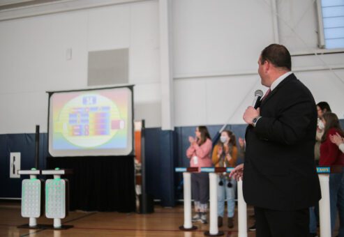 A game show host holding a microphone and looking at a large screen displaying the game board, while participants cheer and clap during the Corporate Feud team building activity in a gymnasium setting.
