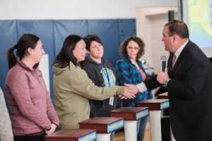 A game show host shaking hands with a participant while holding a microphone, as three other participants look on, during the Corporate Feud team building activity.