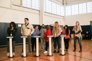A group of participants standing behind individual podiums, ready to compete in the Corporate Feud team building activity, set up in a gymnasium, displaying anticipation and team spirit.