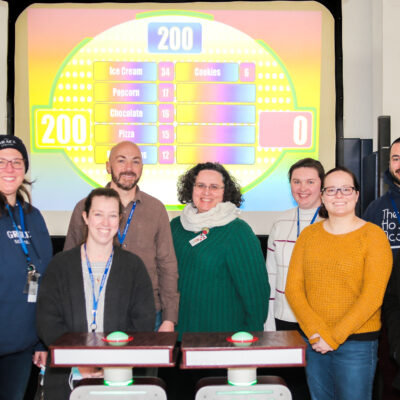 A group of eight participants smiling and standing together in front of a game show setup with a brightly colored game board in the background during the Corporate Feud game.