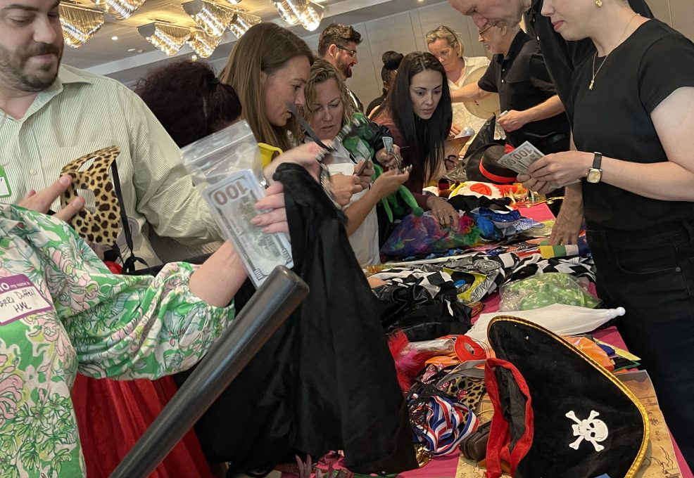 A group of people enthusiastically selecting costumes and props from a table filled with various items like pirate hats, colorful fabrics, and accessories during a team building activity focused on creativity and fun role-playing.