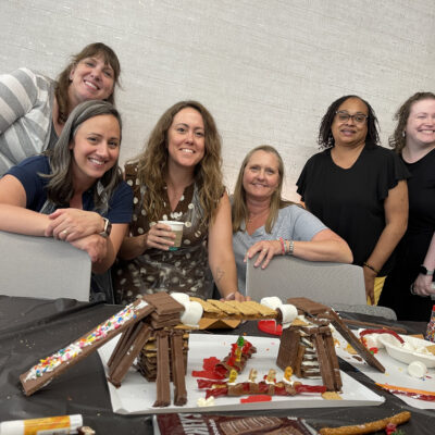 A group of six participants posing proudly behind their chocolate bridge creation during The Chocolate Challenge team building event. The edible bridge is constructed with candy and chocolate materials, showcasing their teamwork and creativity. thumbnail