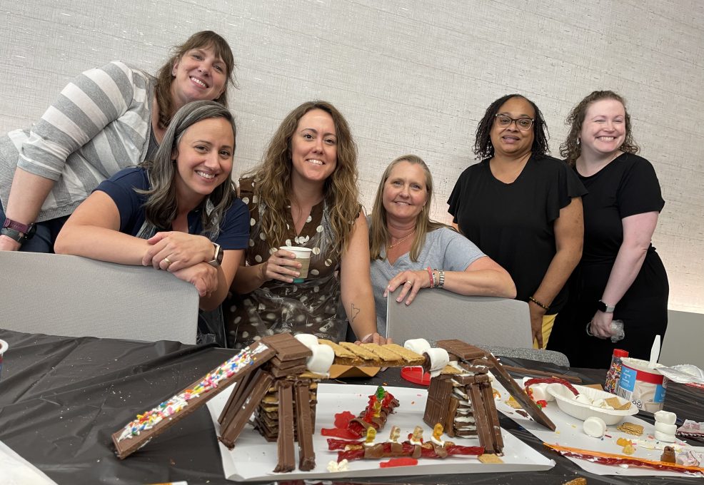 A group of six participants posing proudly behind their chocolate bridge creation during The Chocolate Challenge team building event. The edible bridge is constructed with candy and chocolate materials, showcasing their teamwork and creativity.