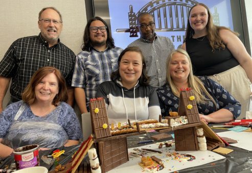 A team of seven poses proudly behind their creative chocolate bridge during The Chocolate Challenge team building event. The structure, made entirely of chocolate, includes detailed elements like wafers and sprinkles. Smiling participants showcase their teamwork and edible engineering skills.