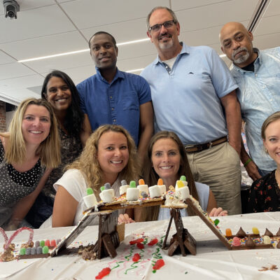 A team proudly poses behind their edible chocolate bridge creation during The Chocolate Challenge team building event. The bridge is decorated with candy, marshmallows, and other sweet treats, highlighting teamwork and creativity. thumbnail