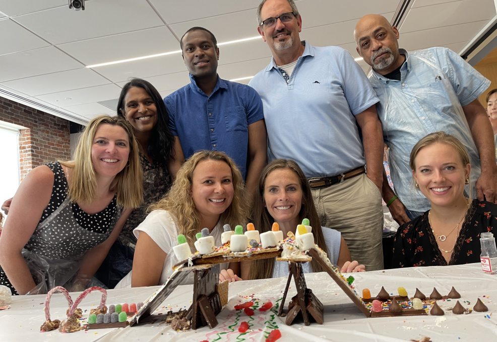 A team proudly poses behind their edible chocolate bridge creation during The Chocolate Challenge team building event. The bridge is decorated with candy, marshmallows, and other sweet treats, highlighting teamwork and creativity.