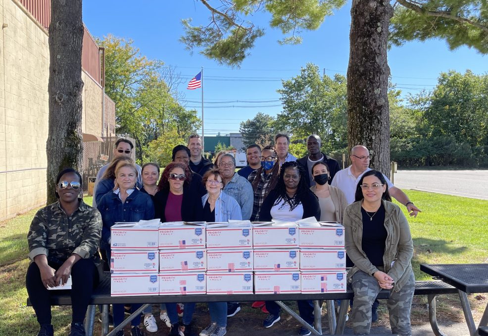 Team members gather with care packages they've assembled during an Operation Military Care team building event, ready to support U.S. military personnel stationed overseas.