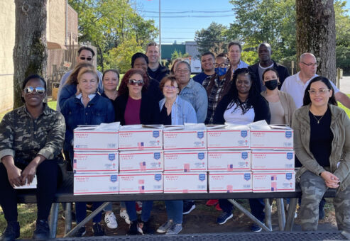 Team members gather with care packages they've assembled during an Operation Military Care team building event, ready to support U.S. military personnel stationed overseas.