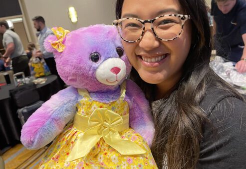 A smiling woman holds a brightly colored purple and pink teddy bear dressed in a yellow dress, as part of a charitable team building event called Team Teddy Rescue Bear.
