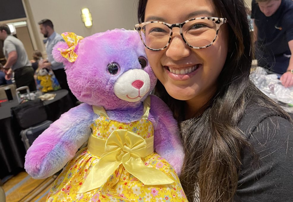 A smiling woman holds a brightly colored purple and pink teddy bear dressed in a yellow dress, as part of a charitable team building event called Team Teddy Rescue Bear.