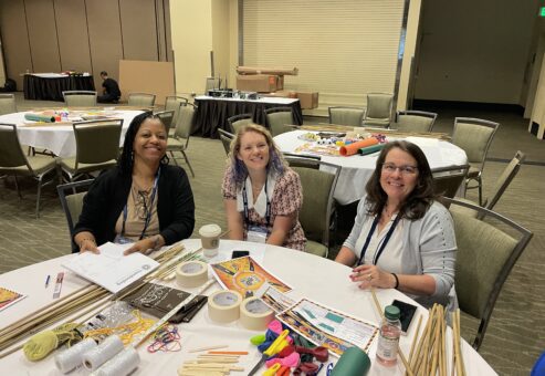 Three participants smile during the Bridging the Divide team building event, seated at a table filled with creative supplies for the bridge-building activity.