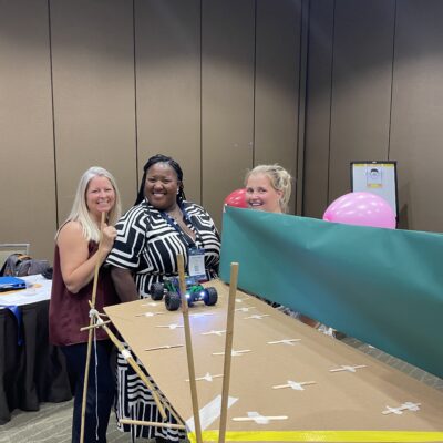 Participants smile next to their completed bridge, ready to test it with a radio-controlled car during the Bridging the Divide team building activity. thumbnail