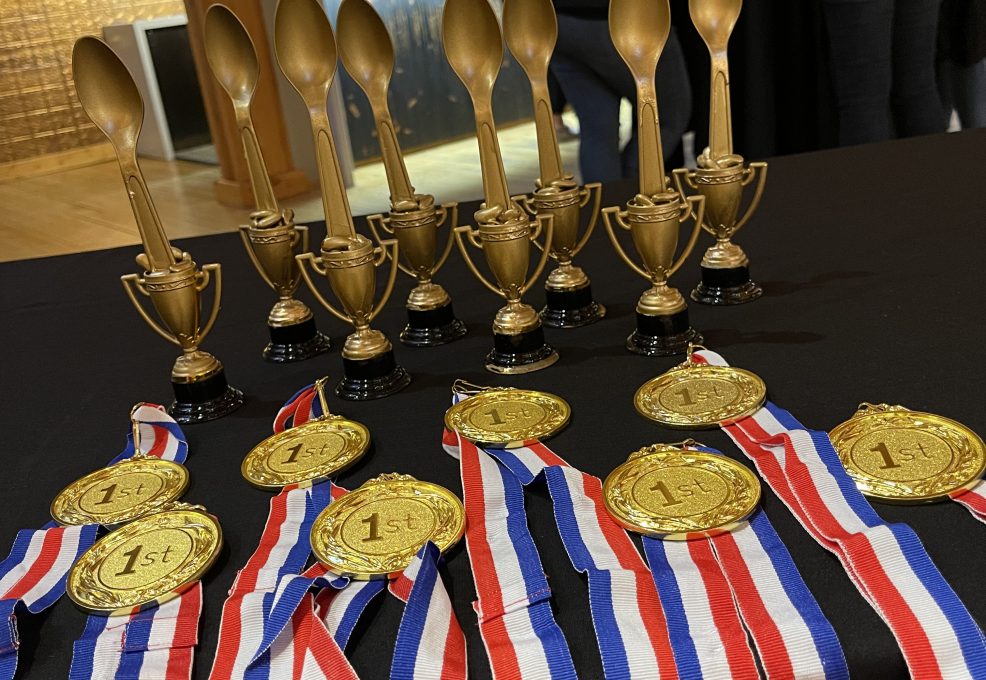 A table displays several gold trophies in the shape of large spoons and medals with '1st' on them. The medals are attached to red, white, and blue ribbons. The trophies and medals are for the culinary team building winners.