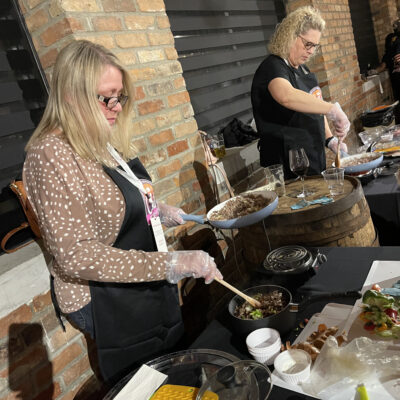 Two women participating in a team building chili cook-off. Both are focused on preparing food at their respective stations. The woman on the left is stirring ingredients in a bowl, wearing a brown polka-dotted cardigan over her apron. The woman on the right is cooking something on a skillet while wearing a black shirt and apron. thumbnail