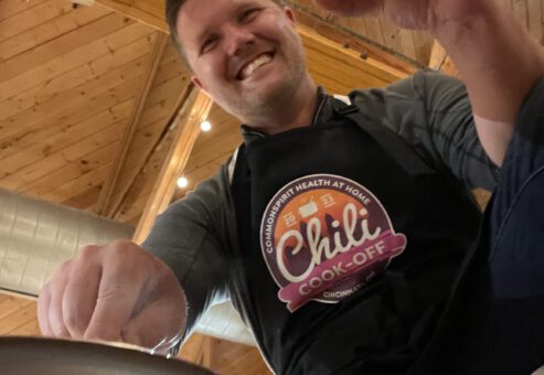 A man is smiling as he participates in a chili cook-off. He is wearing a black apron with the 'Chili Cook-Off' logo prominently displayed. The wooden ceiling and string lights in the background create a cozy atmosphere. The man is holding a glass of beer in one hand and appears to be stirring a pot with the other, fully engaged in the cooking activity.
