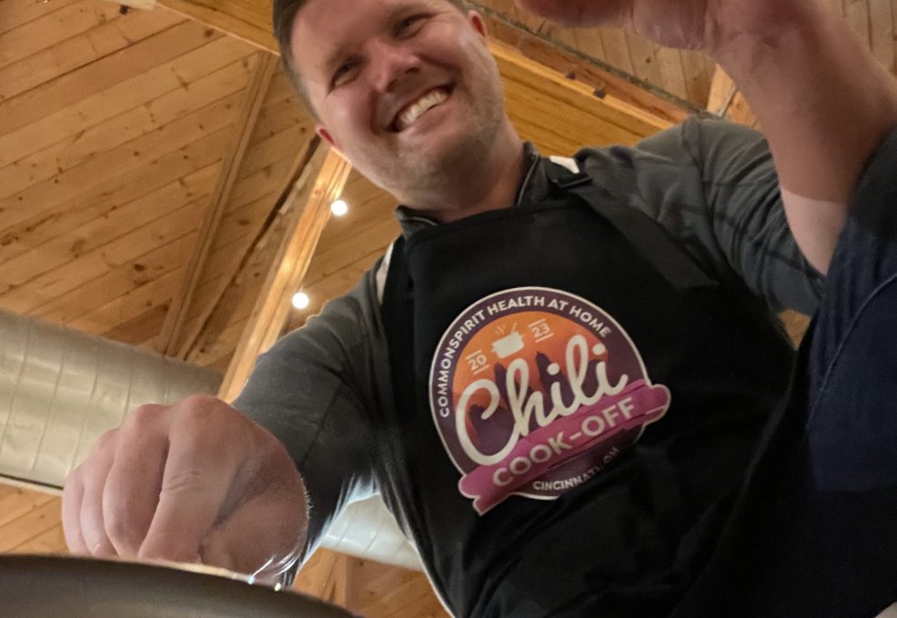 A man is smiling as he participates in a chili cook-off. He is wearing a black apron with the 'Chili Cook-Off' logo prominently displayed. The wooden ceiling and string lights in the background create a cozy atmosphere. The man is holding a glass of beer in one hand and appears to be stirring a pot with the other, fully engaged in the cooking activity.