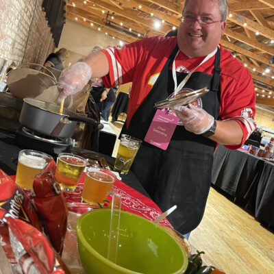 A man wearing a red sports jersey and a black apron is smiling while stirring a pot on a stove during a culinary team building event. He holds a set of tongs in one hand and is wearing plastic gloves. In front of him are cups of beer, a bag of Doritos chips, and a green bowl. thumbnail