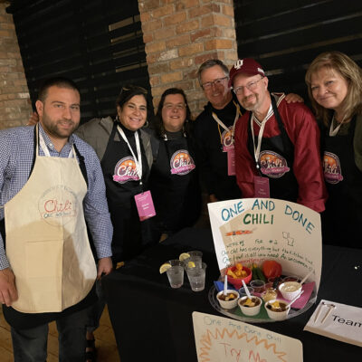 A group of six participants posing at a team building chili cook-off event behind their table. The table displays a sign reading 'One and Done Chili' with a description and a tray of chili servings garnished with toppings. The participants are smiling and wearing 'Chili Cook-Off' aprons. thumbnail