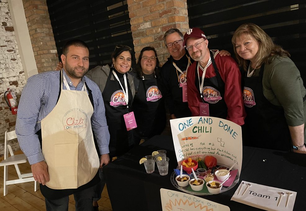 A group of six participants posing at a team building chili cook-off event behind their table. The table displays a sign reading 'One and Done Chili' with a description and a tray of chili servings garnished with toppings. The participants are smiling and wearing 'Chili Cook-Off' aprons.